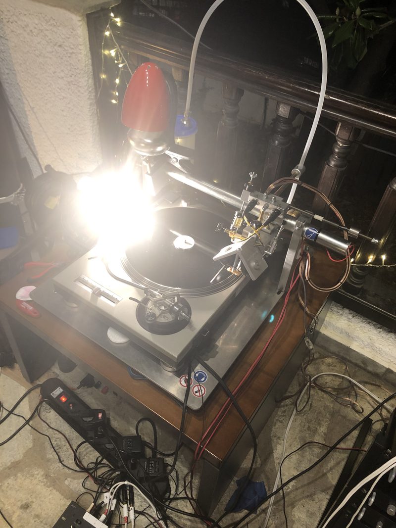 A cutting lathe mounted above a turntable, its cutting head poised over a blank disc under a desk lamp, with wiring and a suction tube running to one side.